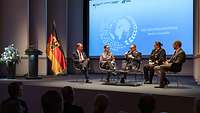 fünf Personen auf einer kleinen Konzertbühne während einer Podiumsdiskussion neben Rednerpult und Deutschlandflagge